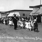 Anchorage elementary school students marching in a parade, Anchorage, 1915 or 1916.  The woman in the dark clothing walking alongside them has been identified as Orah Dee Clark.  