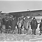 A group of men, probably pilots or employees of Star Airlines, with one of the company airplanes. Most of the men wear Alaska Native cold weather clothing, including reindeer fur parkas, reindeer or seal fur mukluks, and moosehide gauntlets.   