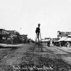 A look down Anchorage’s Fourth Avenue, the main business district, with a photographer preparing to shoot an elevated view of the street in about 1916.  A sign for “A.A. Shonbeck, General Merchandise” can be seen on the right side of the street.  Shonbeck sold a wide range of merchandise, including cars, miners’s supplies, fuel, and hay.
